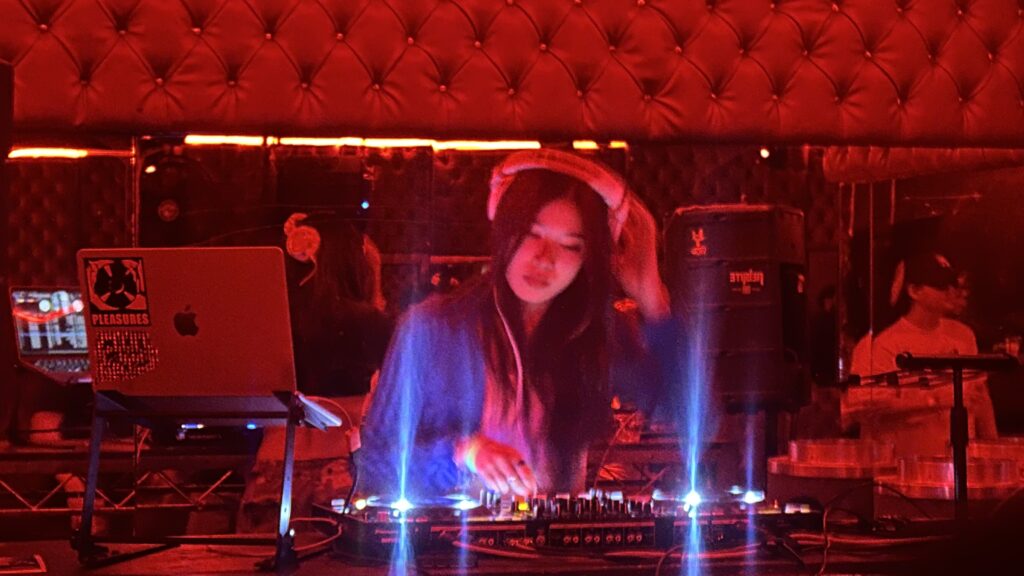 DJ Ema performing in a dimly lit venue with red lighting, focused on her equipment.